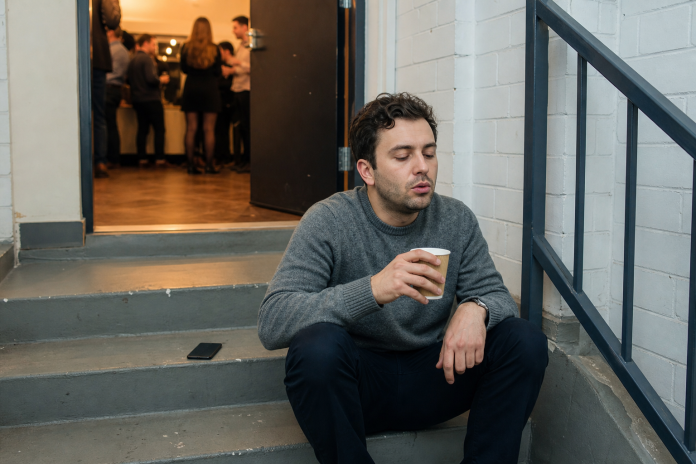 Man sitting alone on a stairwell landing with a cup of tea, taking a break from a noisy office party behind the door