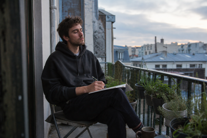 Morning reflection on a balcony Young man on an apartment balcony at dawn writing in a notebook with his eyes closed