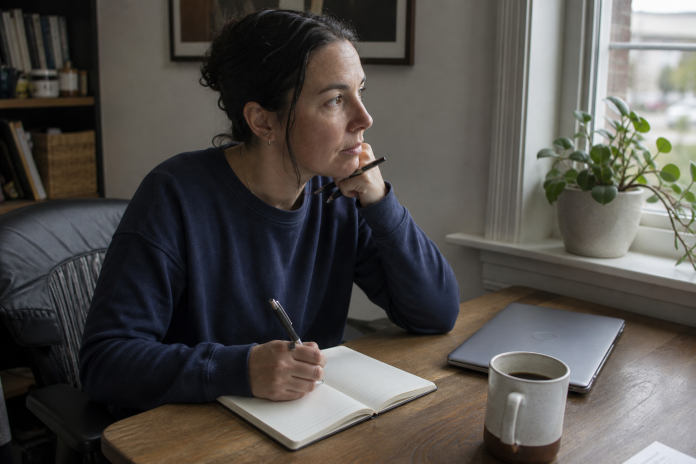 Woman at a desk by a window writing in a notebook with two columns, pen paused in thought
