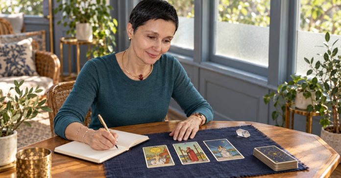 Woman with short black hair writing in a notebook beside three tarot cards on an indigo mat.