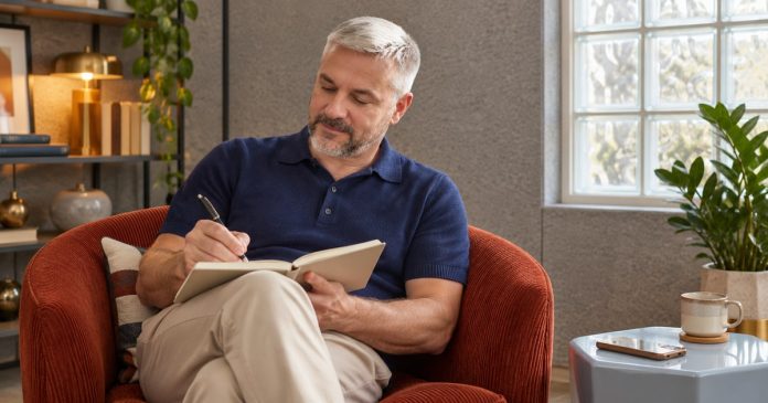 Man writing in a notebook on his lap with a phone set aside on a pale-blue side table