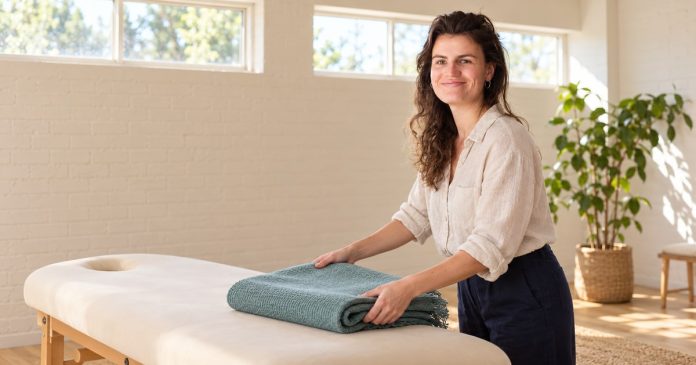 Woman arranging a folded teal blanket on an empty treatment couch in a bright room with high windows